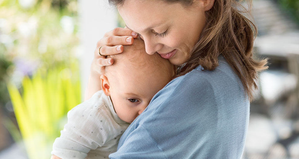 A woman in a blue shirt is holding a baby with a white outfit, both facing the camera.