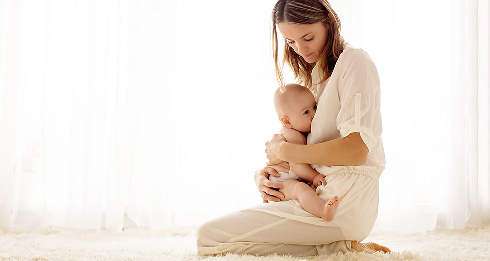 A woman in white clothing is holding a baby on a white carpeted floor, with a white curtain in the background.