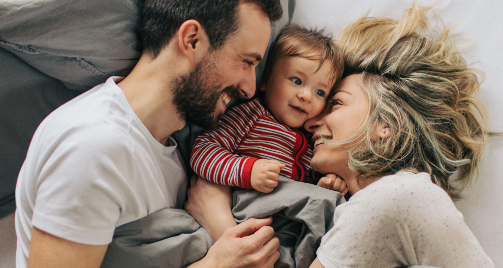 Three people, two adults and a child, are lying on a bed, looking at each other.