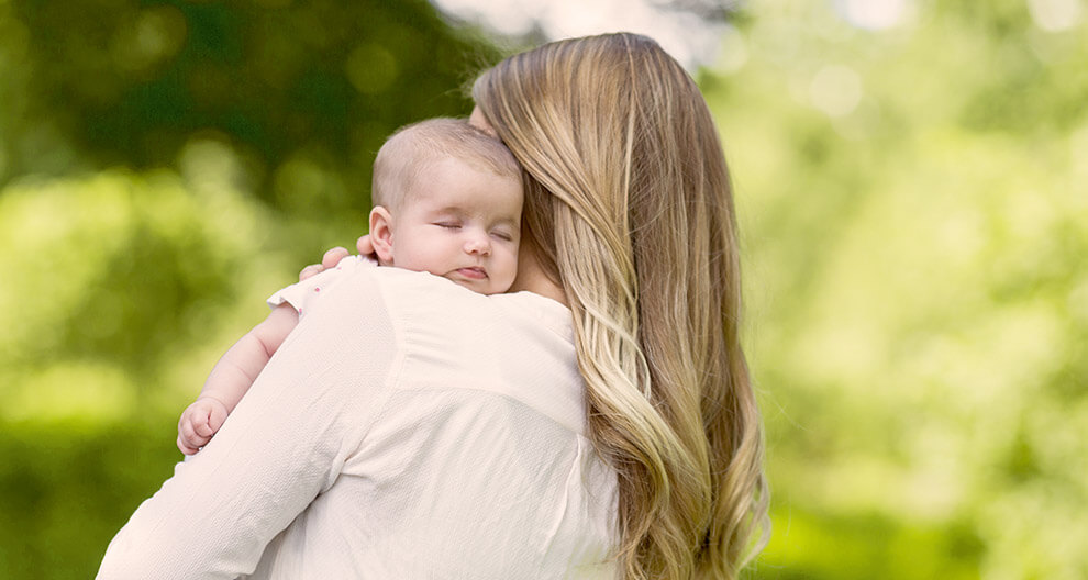 A woman with long hair holds a sleeping baby against a blurred green background.