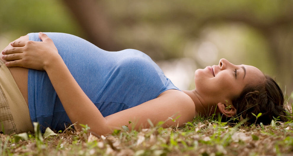A pregnant woman lies on grass, her hand resting on her belly.
