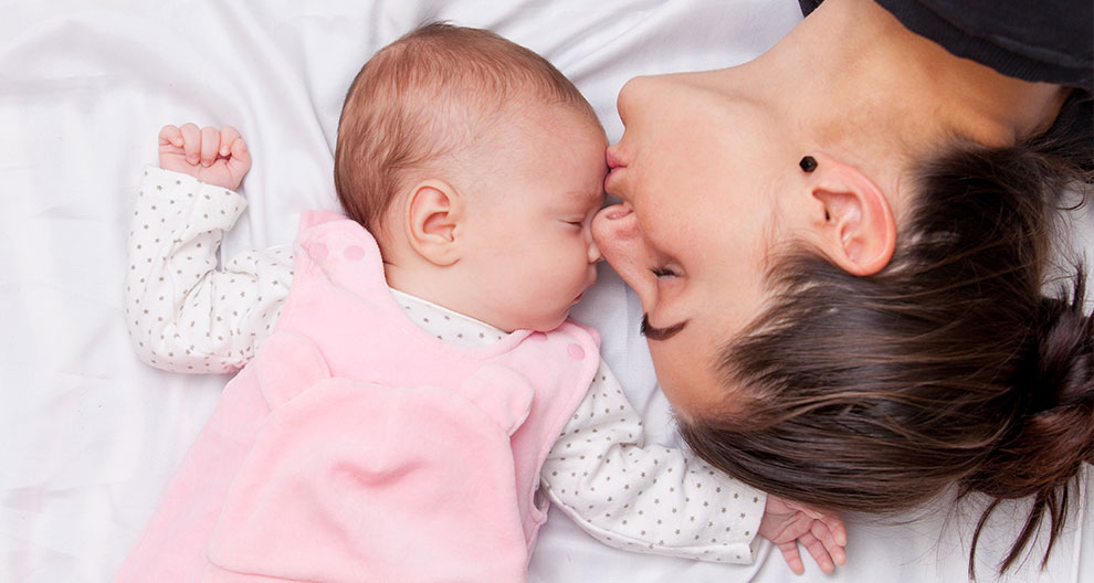 A woman and a baby lie on a white surface, with the woman leaning over to kiss the baby's forehead.