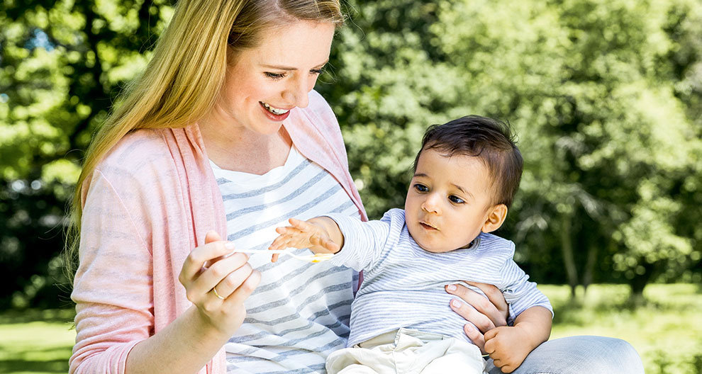 A woman in a pink cardigan and striped shirt is feeding a baby in a striped shirt and white pants with a spoon in a park with green trees in the background.