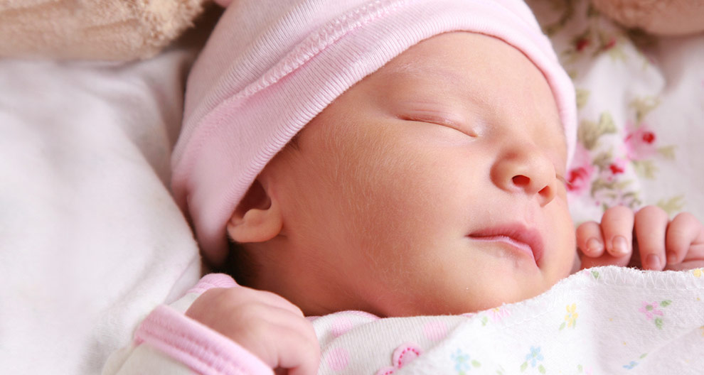 A close-up of a sleeping baby's face, wearing a pink hat and swaddled in a white blanket with colorful floral patterns.