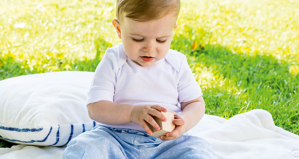A baby sits on a blanket outdoors, holding a wooden block.