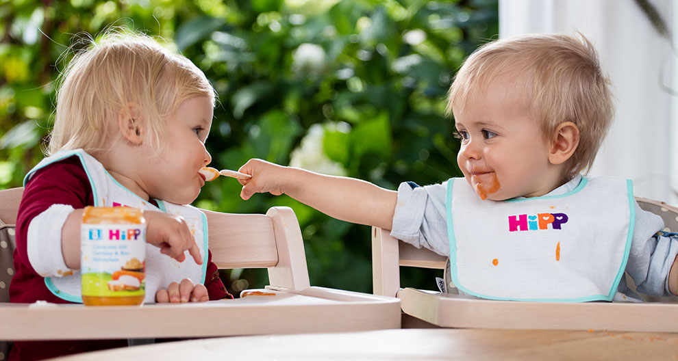 Two toddlers are seated at a table, feeding each other with food from a jar.