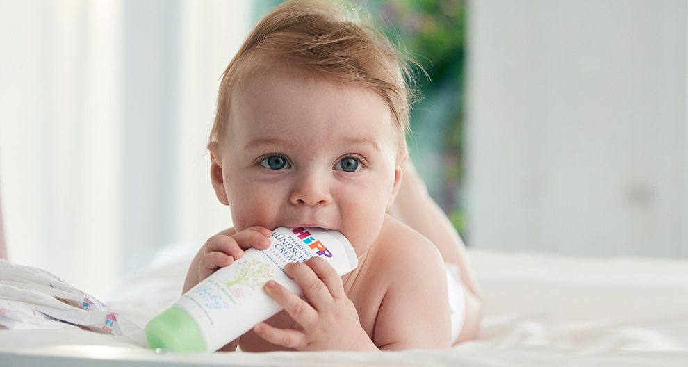 A baby with light hair and blue eyes is biting a green and white tube.