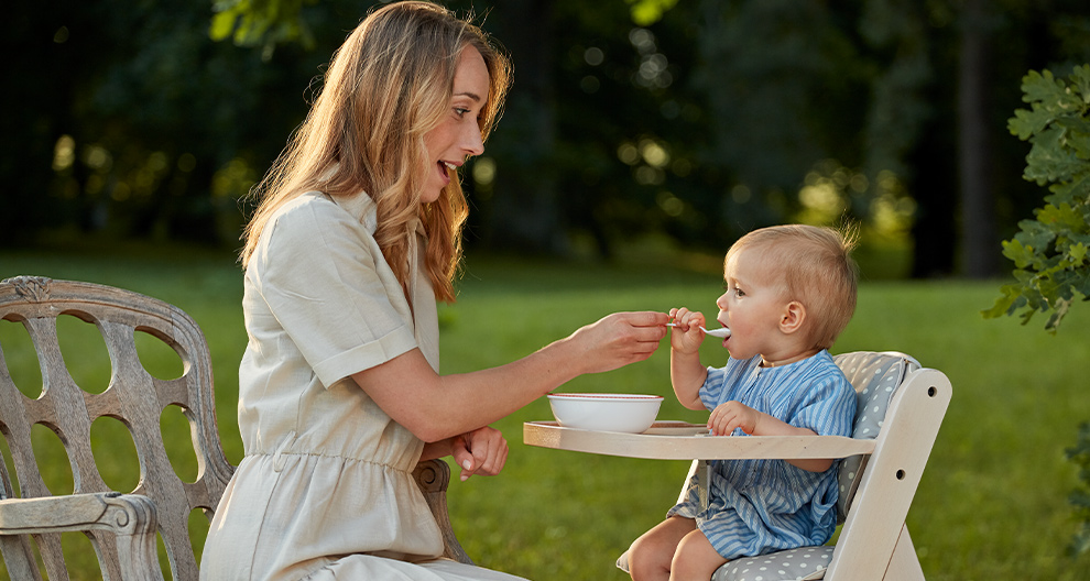 A woman in a white dress is feeding a baby seated in a high chair outdoors.