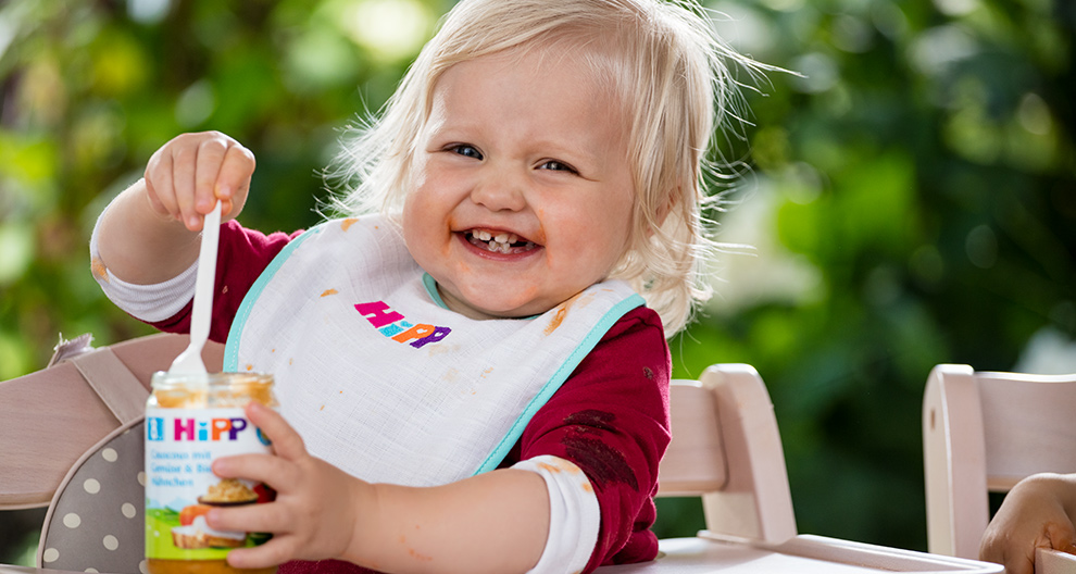 A toddler with blonde hair is seated outdoors, wearing a bib, and holding a jar of food.