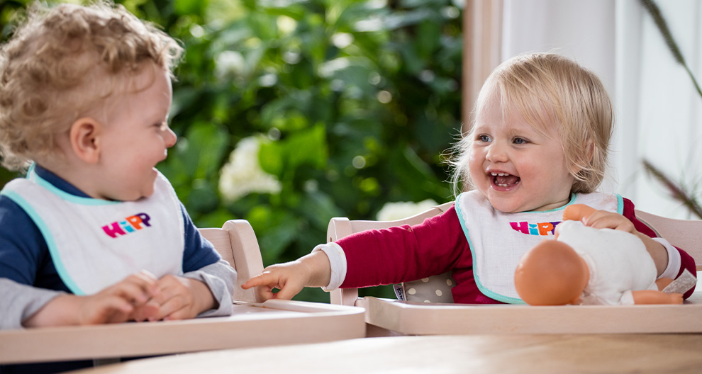 Two toddlers sit in high chairs, one wearing a blue shirt and bib with the word "HIP" and the other in a red shirt and bib with the same word, both looking at each other with a doll on the table between them.