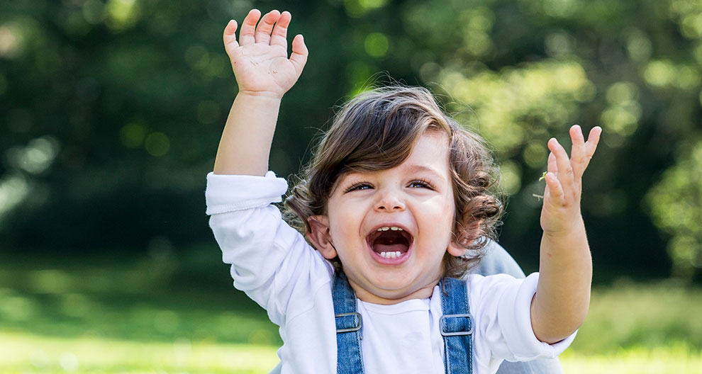 A child with curly hair, wearing a white shirt and blue suspenders, is raising both hands and has an open mouth, possibly shouting or laughing.