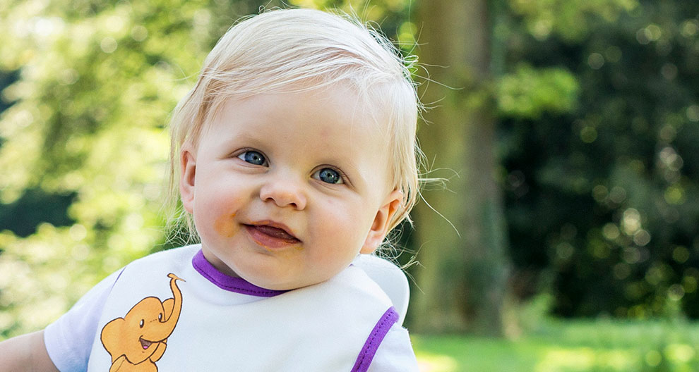 A young child with light hair and blue eyes is wearing a white bib with a purple trim and a cartoon elephant design, standing outdoors with a blurred green background.