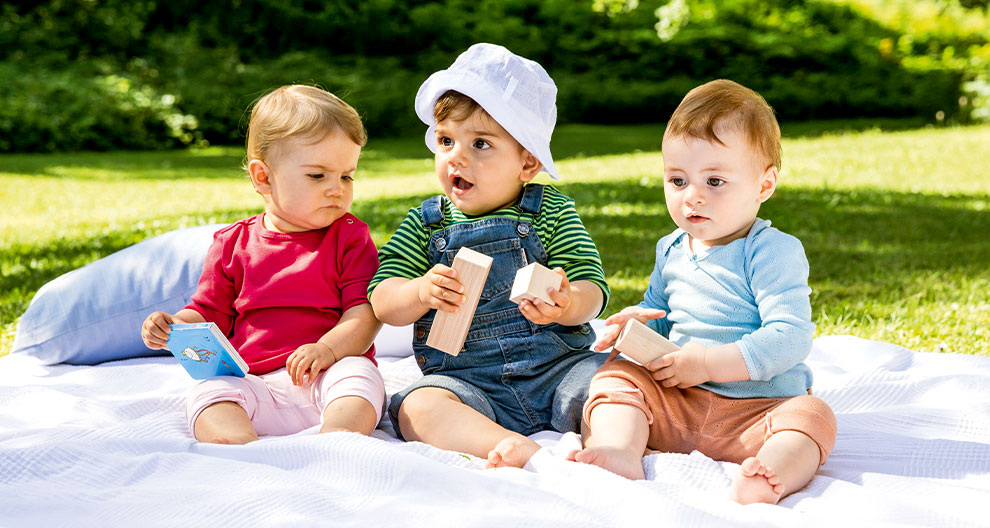 Three toddlers sit on a blanket outdoors, each holding a toy.