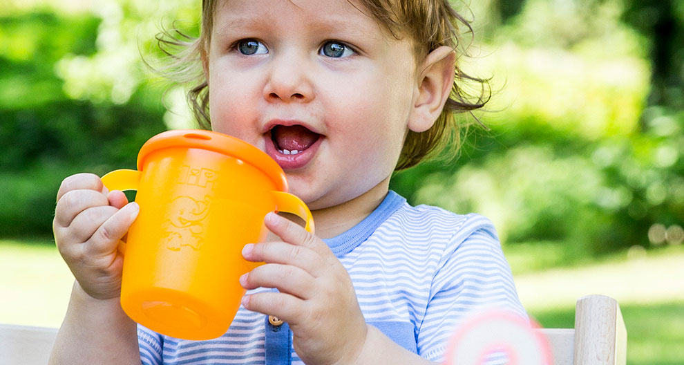 A toddler with curly hair is drinking from an orange cup with a cartoon character embossed on it.