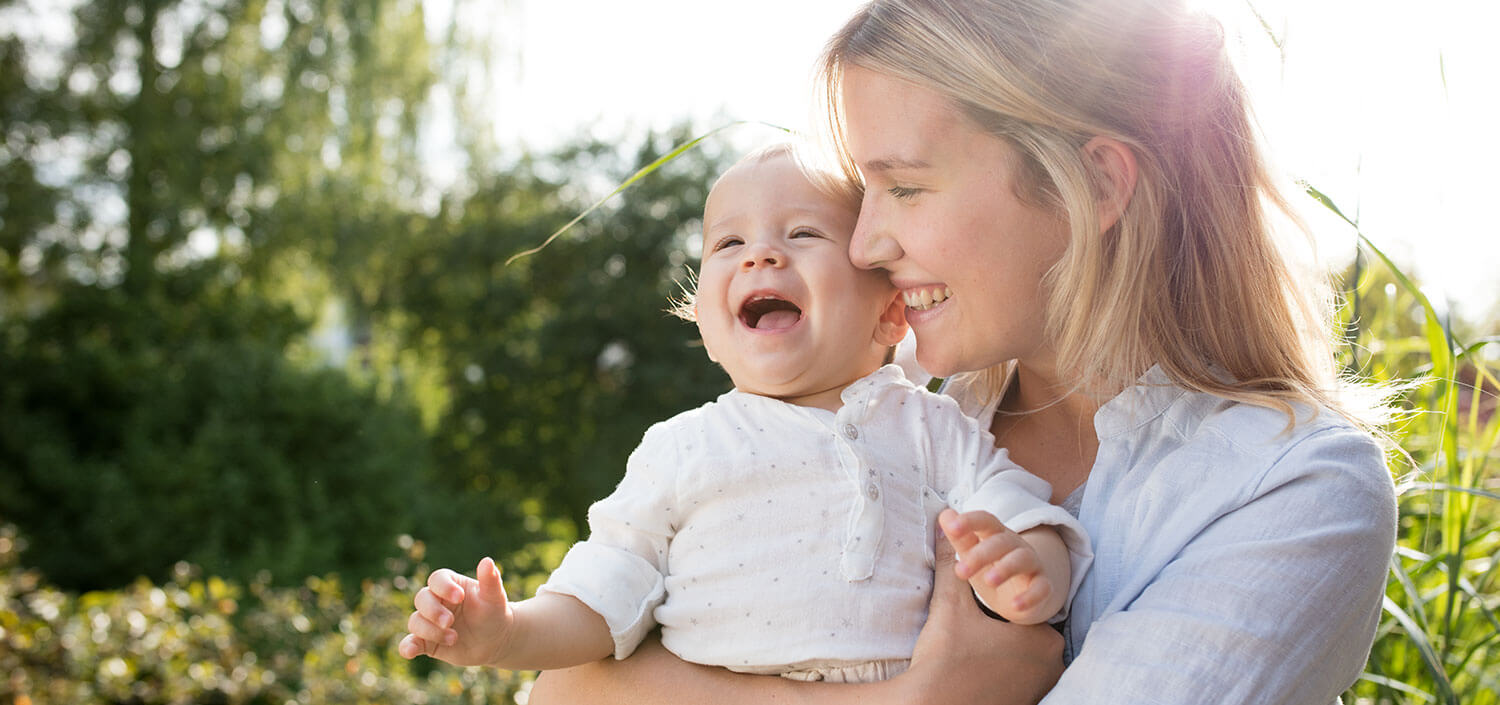 A woman holds a laughing baby outdoors.