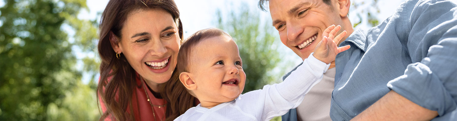 Three people outdoors, two adults and a baby, looking at each other.