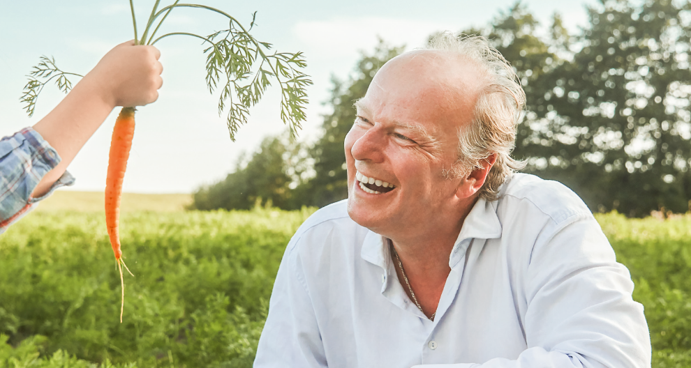 An elderly man, wearing a white shirt, is smiling as he looks at a child's hand presenting a freshly picked carrot with its green leaves.