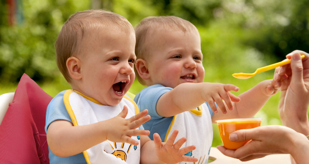 Dvije bebe u podbradnjacima posežu za hranom žlicom.