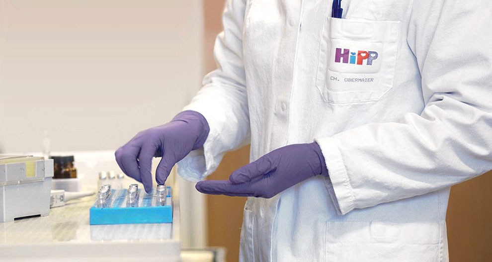 Osoba u bijelom laboratorijskom ogrtaču s logotipom "HiPP" i imenom "CH. OBERMAIER" nosi ljubičaste rukavice i rukuje plavim stalkom s malim posudama.