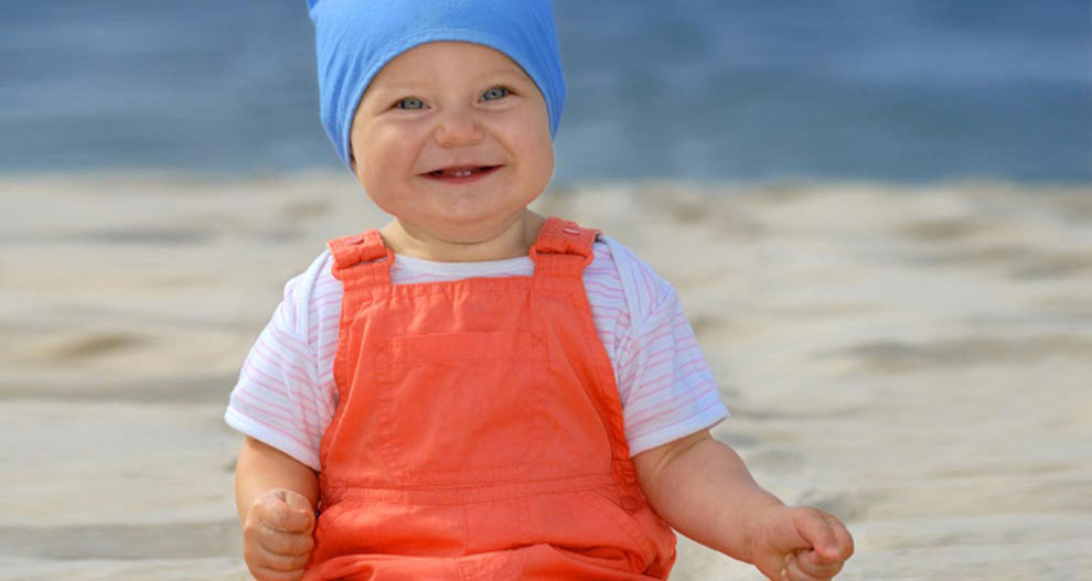 Beba sjedi na pješčanoj plaži, odjevena u plavu kapu i narančasti kombinezon.