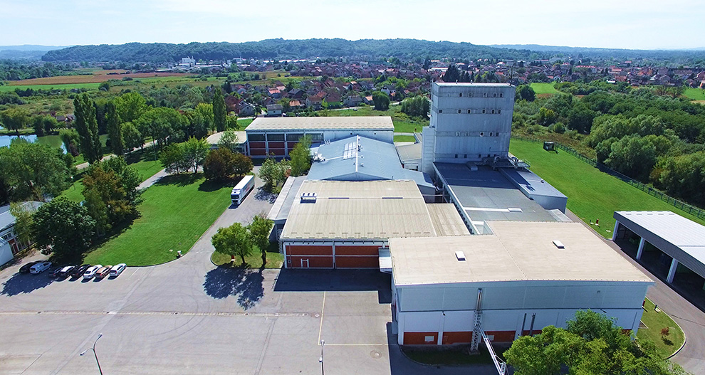 Zračni pogled na kompleks s velikom zgradom, okružen zelenilom, cestama i parkirnim mjestima, s gradom i brdima u pozadini.