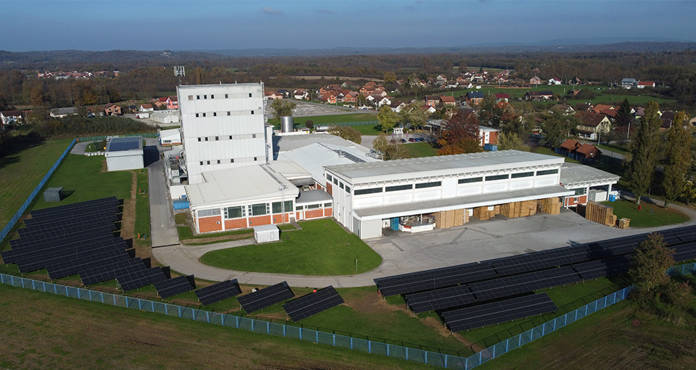 Zračni pogled na kompleks zgrada s velikom instalacijom solarnih panela, okružen plavom ogradom, smješten nasuprot stambenog područja i zelenih polja u pozadini.