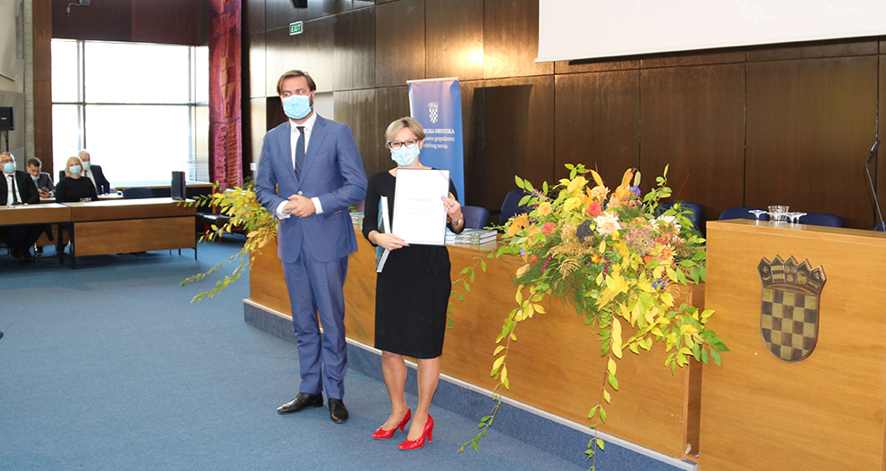 Dvije osobe stoje u formalnom okruženju, jedna drži certifikat, sa štitom vidljivim na podiju.